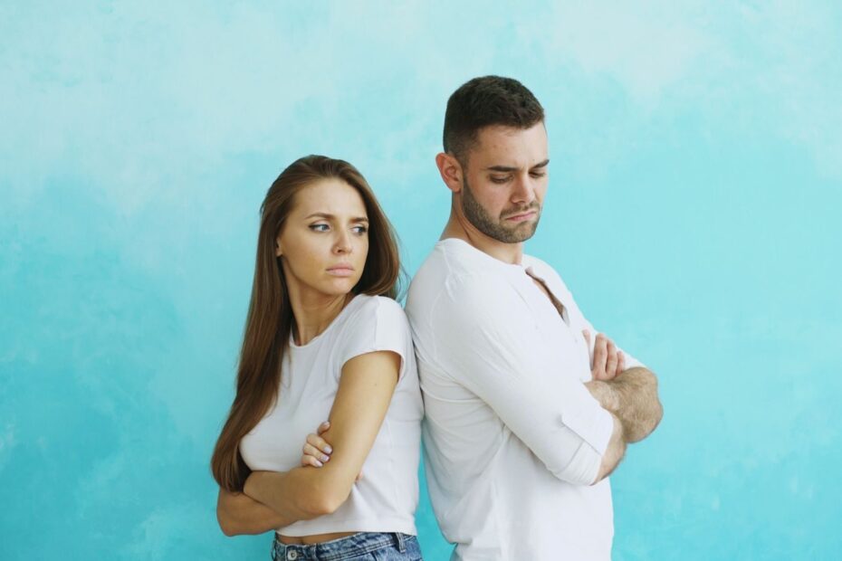 A tense couple stands back to back with crossed arms against a blue background.